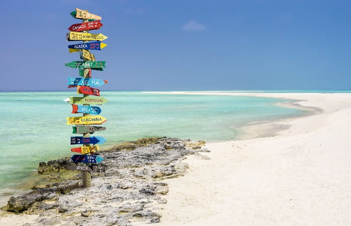 Panneau coloré indiquant différentes destinations touristiques planté sur une plage de sable blanc aux eaux turquoise.
