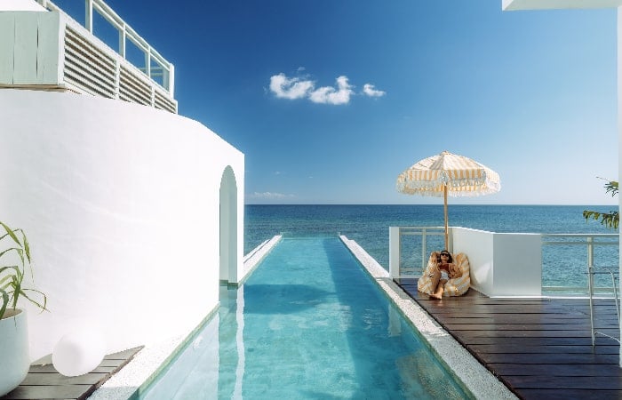 Femme assise sous un parasol au bord d'une piscine à débordement donnant sur la mer, dans un cadre architectural blanc et épuré.