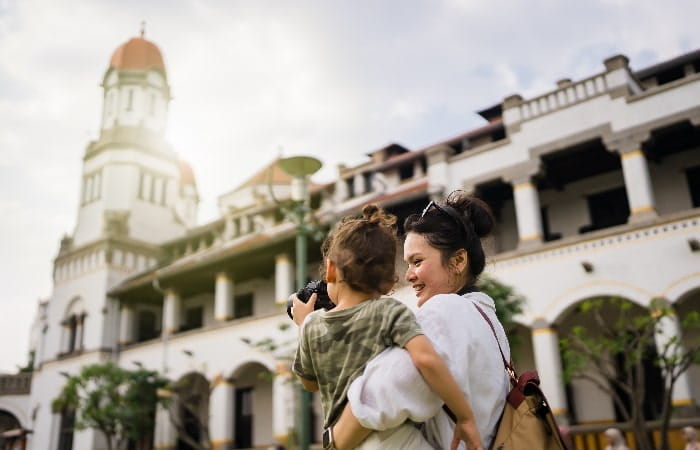 Mère souriante portant son enfant qui tient un appareil photo, devant un bâtiment historique au style colonial.