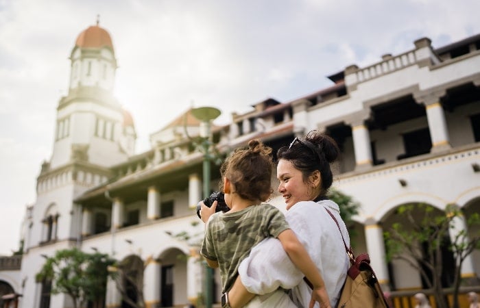 Mère souriante portant son enfant qui tient un appareil photo, devant un bâtiment historique au style colonial.