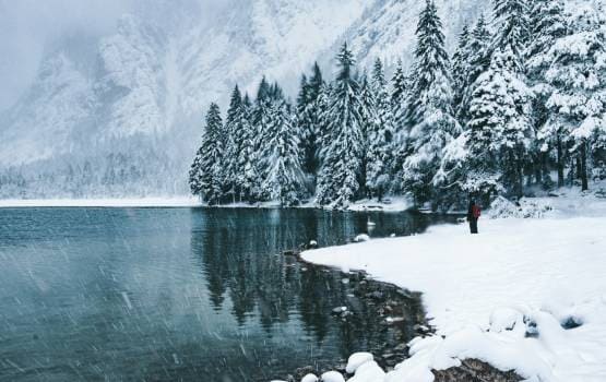 Lac bordé d’une forêt enneigée avec montagnes en arrière-plan et chute de neige légère.