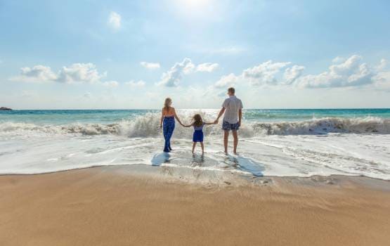 Trois personnes se tenant la main au bord de l’océan, face aux vagues, sur une plage de sable sous un ciel ensoleillé.