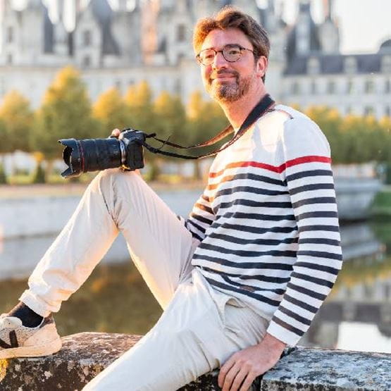 Un homme avec son genoux posé sur un muret devant des chateaux et un appareil photo à la main. 