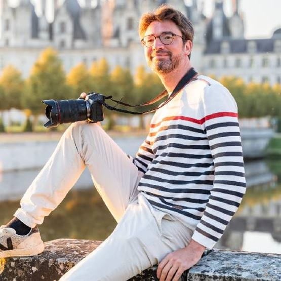 Un homme avec son genoux posé sur un muret devant des chateaux et un appareil photo à la main.