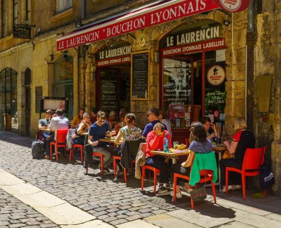 Terrasse de bouchon lyonnais avec clients attablés dans une rue pavée ensoleillée.